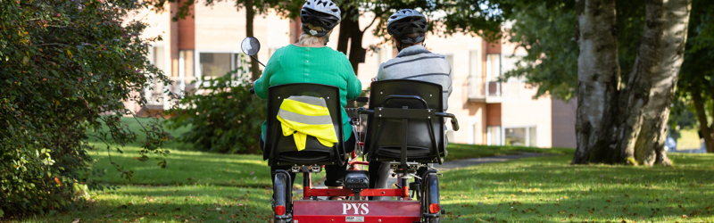 To personer på dobbeltcykel med en ældre- eller handicapbolig i baggrunden